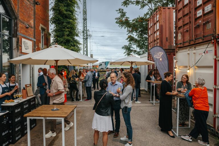Crowd of people at an outdoor social event under white umbrellas between a brick building and shipping containers, with drinks and food nearby.
