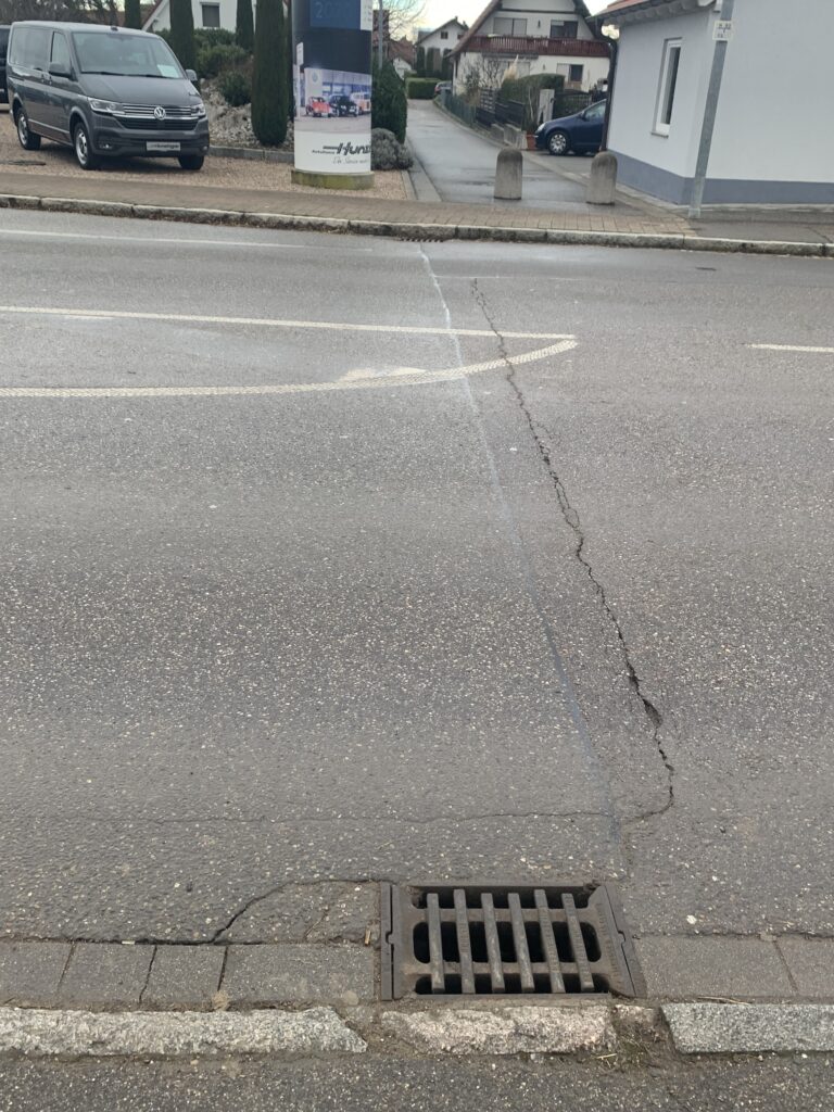 Storm drain grate in asphalt at the curb with cracked pavement and a residential street in the background.