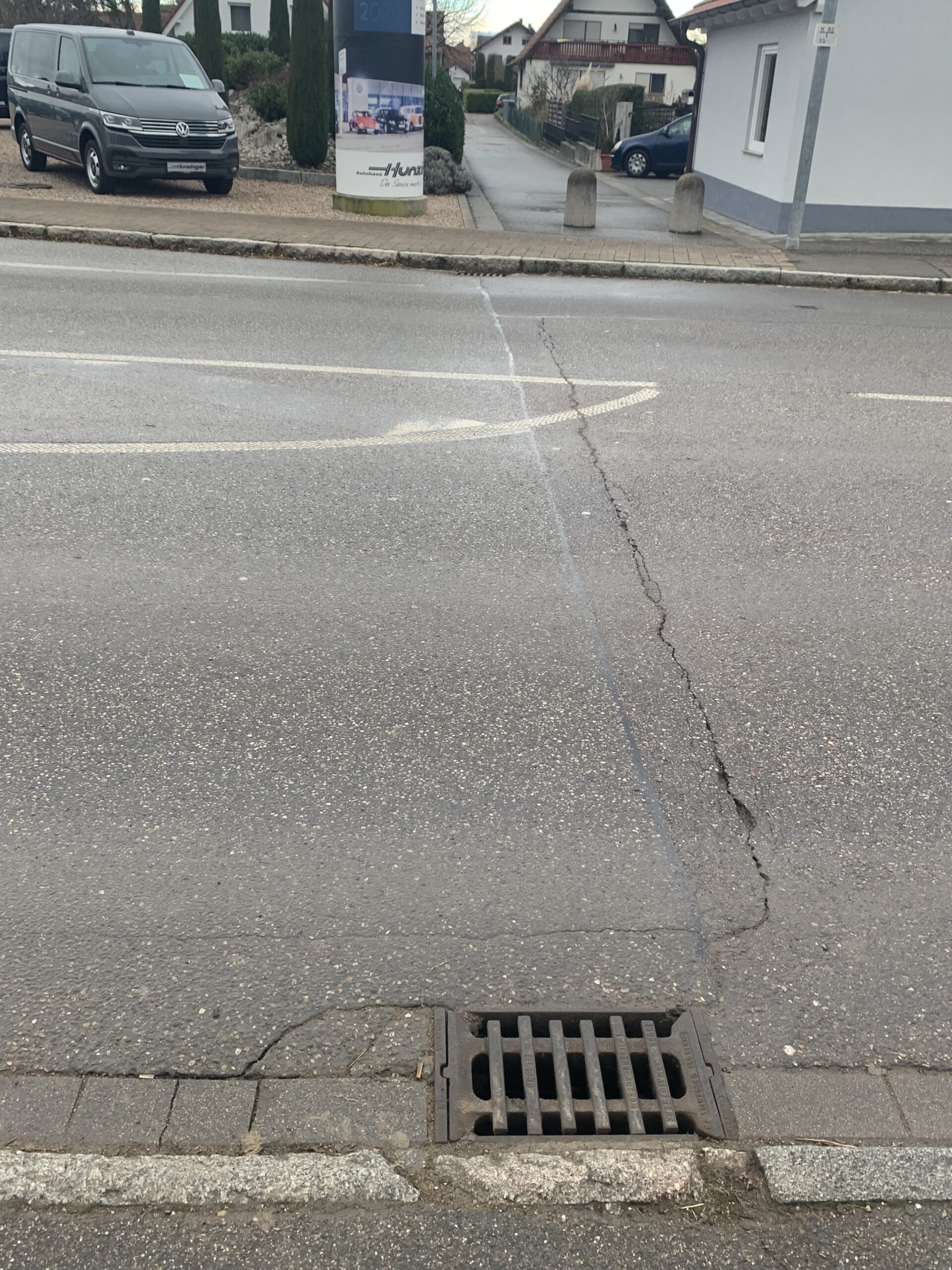 Storm drain grate in asphalt at the curb with cracked pavement and a residential street in the background.