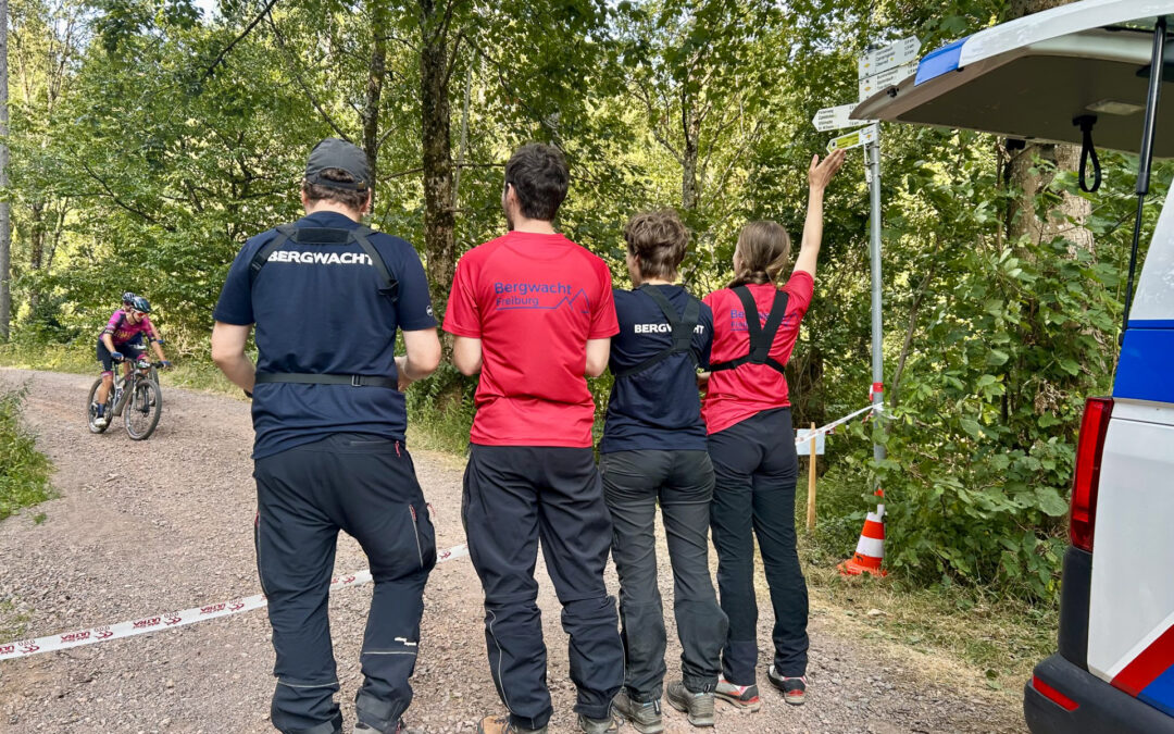 Rescue team members in red and blue shirts stand on a gravel forest trail, a cyclist visible in the background near a signpost and van.
