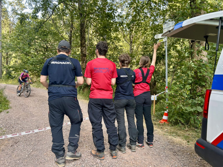 Rescue team members in red and blue shirts stand on a gravel forest trail, a cyclist visible in the background near a signpost and van.