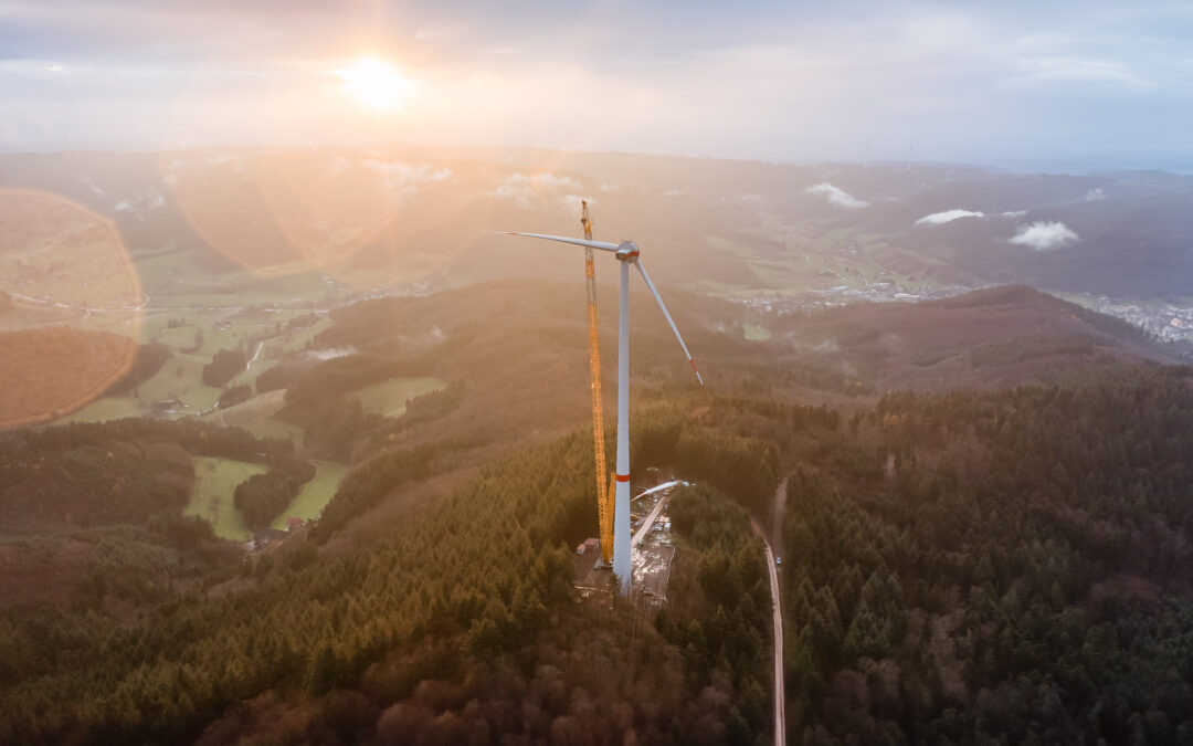 Aerial view of a wind turbine on a hill with a crane nearby and a warm sunset glow over a forested valley.