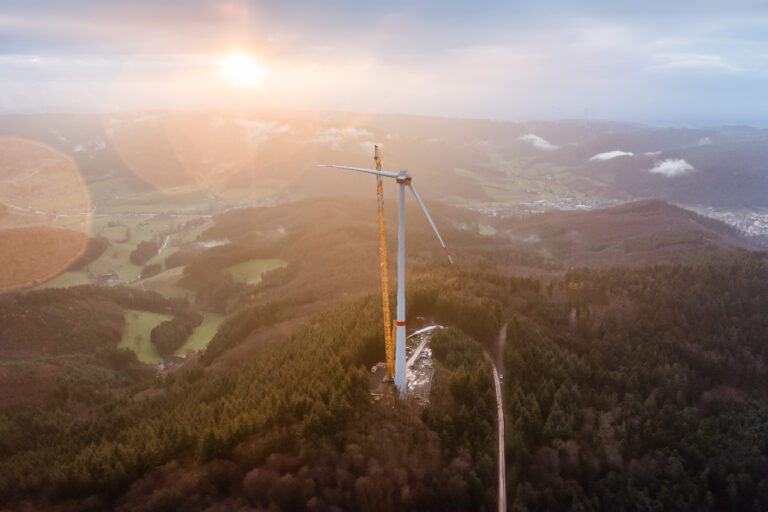 Aerial view of a wind turbine on a hill with a crane nearby and a warm sunset glow over a forested valley.