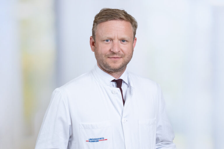 Portrait of a male doctor in a white coat, looking at the camera in a bright clinical setting.