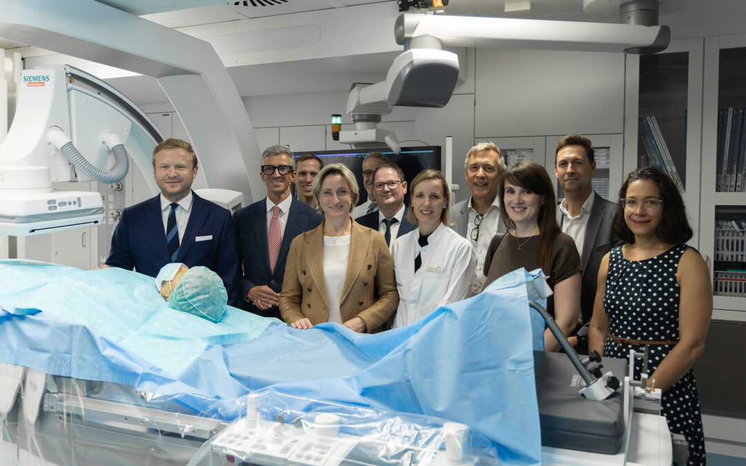 Group of doctors and staff in a modern operating room with a patient on an operating table covered in blue drapes and Siemens imaging equipment nearby.