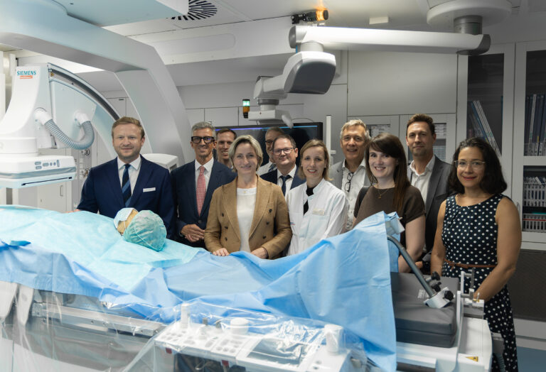 Group of doctors and staff in a modern operating room with a patient on an operating table covered in blue drapes and Siemens imaging equipment nearby.
