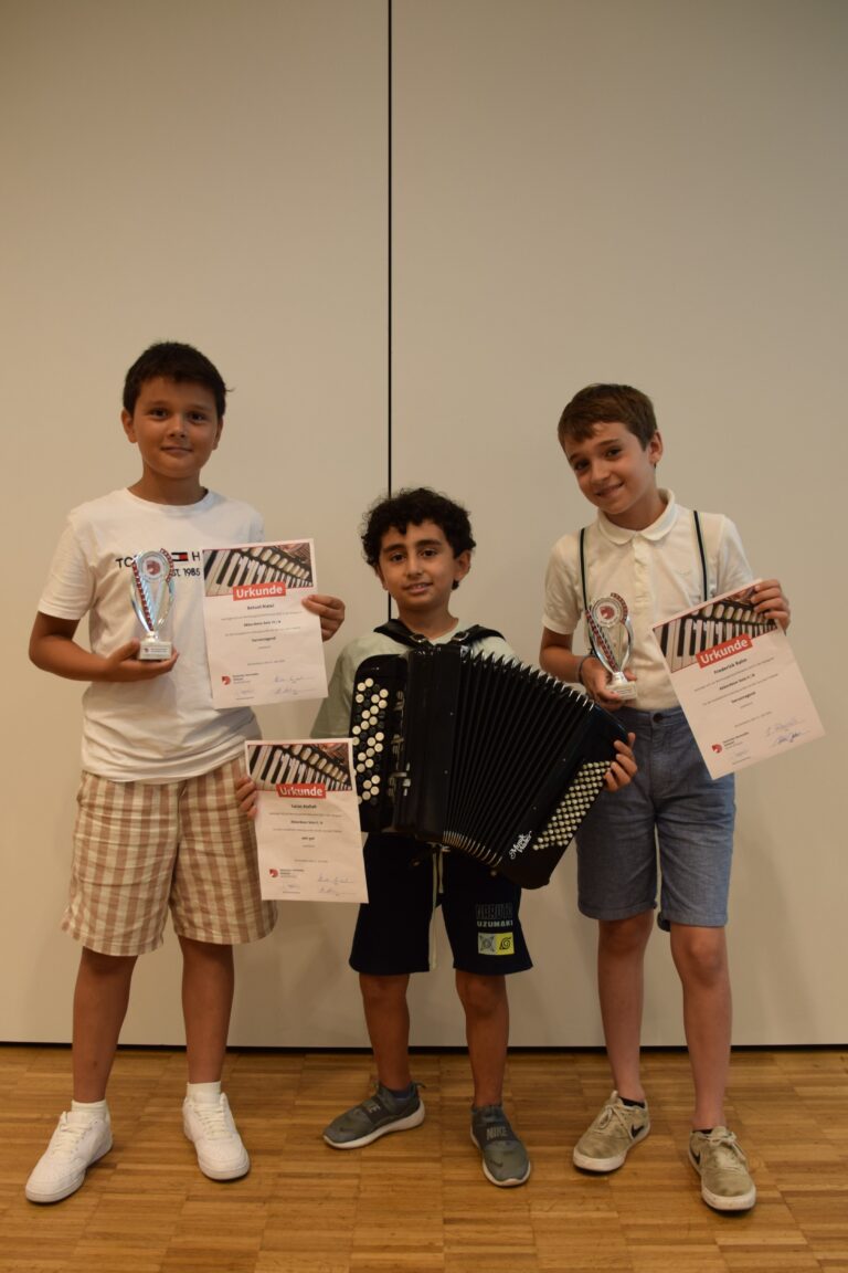 Three boys pose with awards and certificates, one holding a trophy; an accordion rests in front of them against a plain wall background.