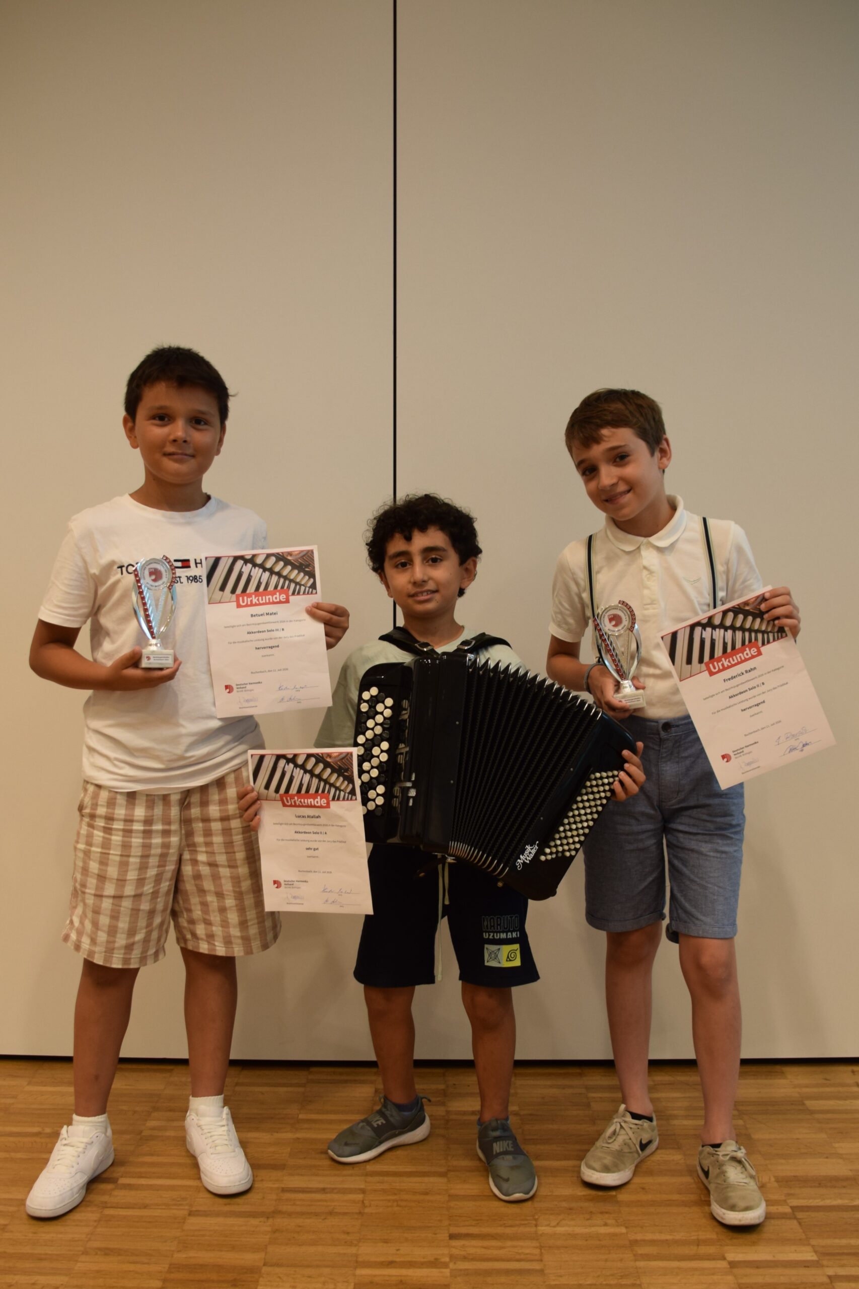 Three boys pose with awards and certificates, one holding a trophy; an accordion rests in front of them against a plain wall background.