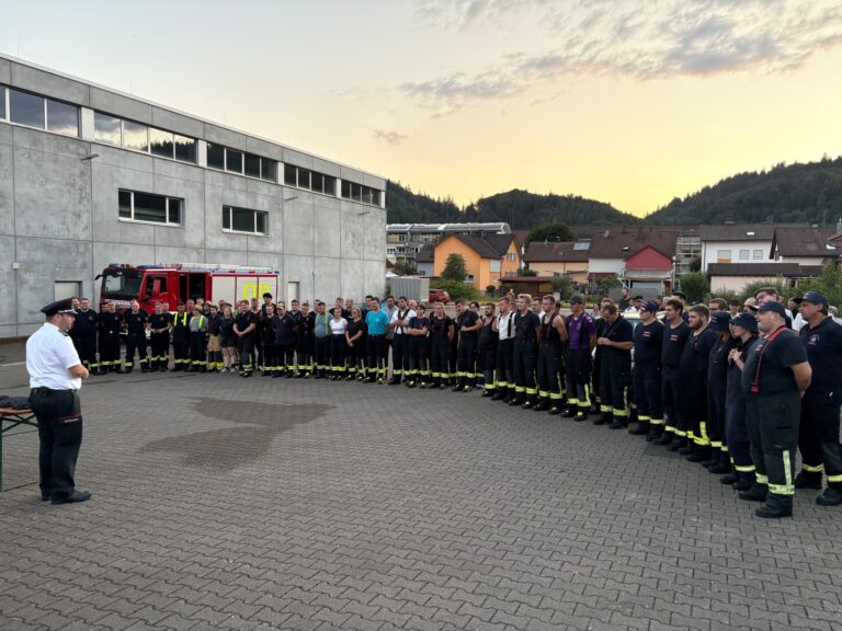 Group of firefighters in uniform standing in formation outside a building with a red fire truck nearby at sunset.