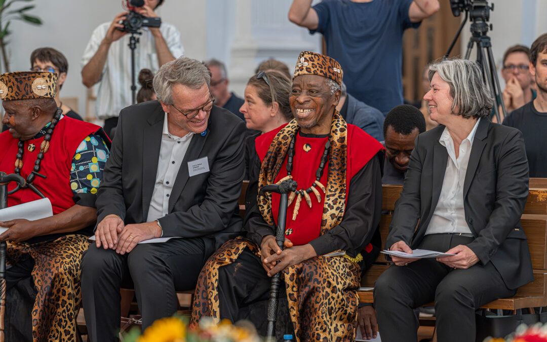 Group of adults seated on a wooden bench; two wear black suits and the others wear red and leopard-print traditional outfits with bead necklaces, at a ceremony with camera crew in the background.
