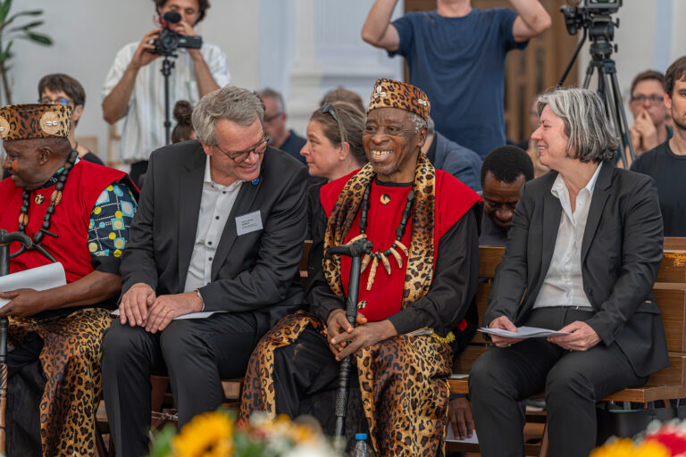 Group of adults seated on a wooden bench; two wear black suits and the others wear red and leopard-print traditional outfits with bead necklaces, at a ceremony with camera crew in the background.