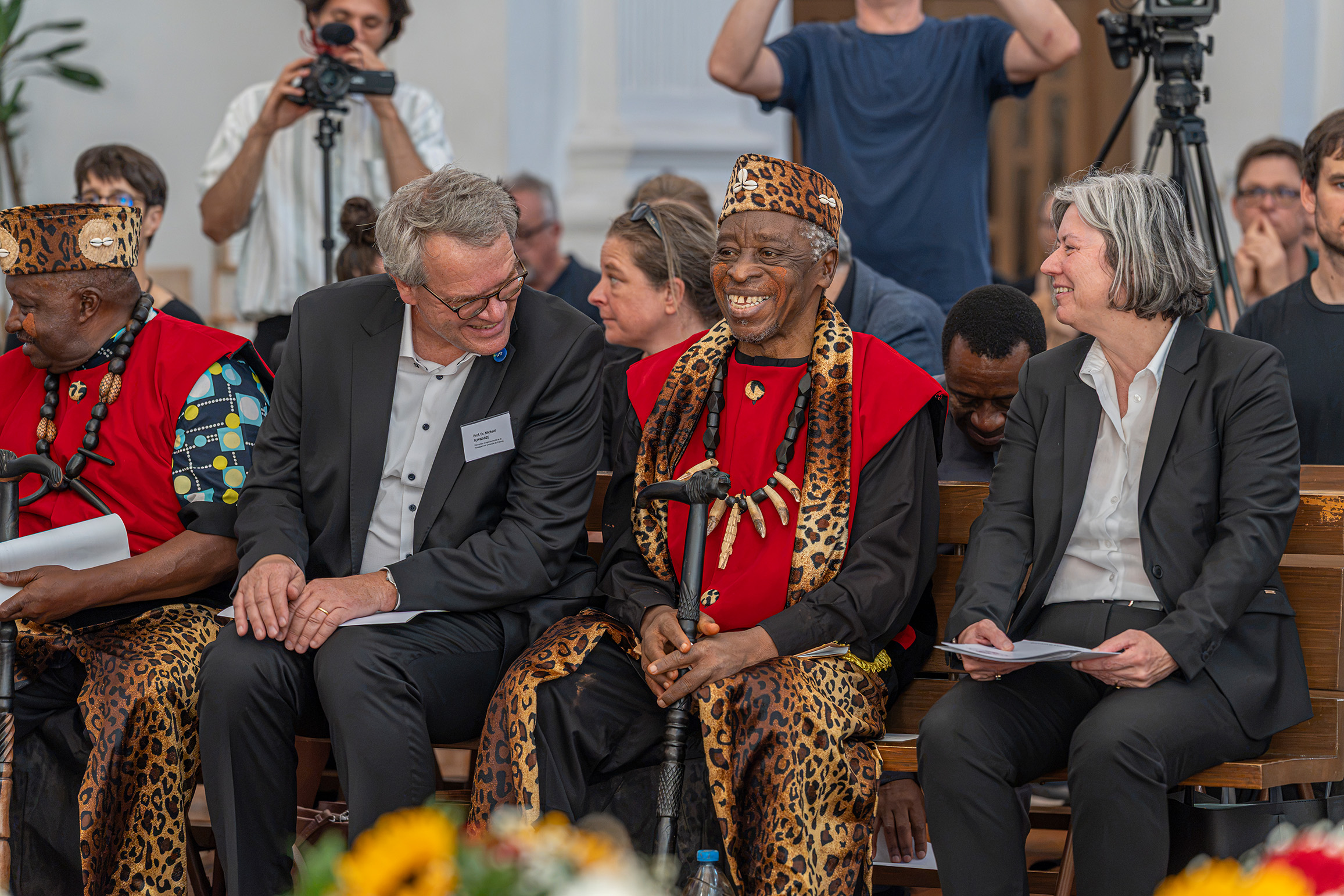 Group of adults seated on a wooden bench; two wear black suits and the others wear red and leopard-print traditional outfits with bead necklaces, at a ceremony with camera crew in the background.