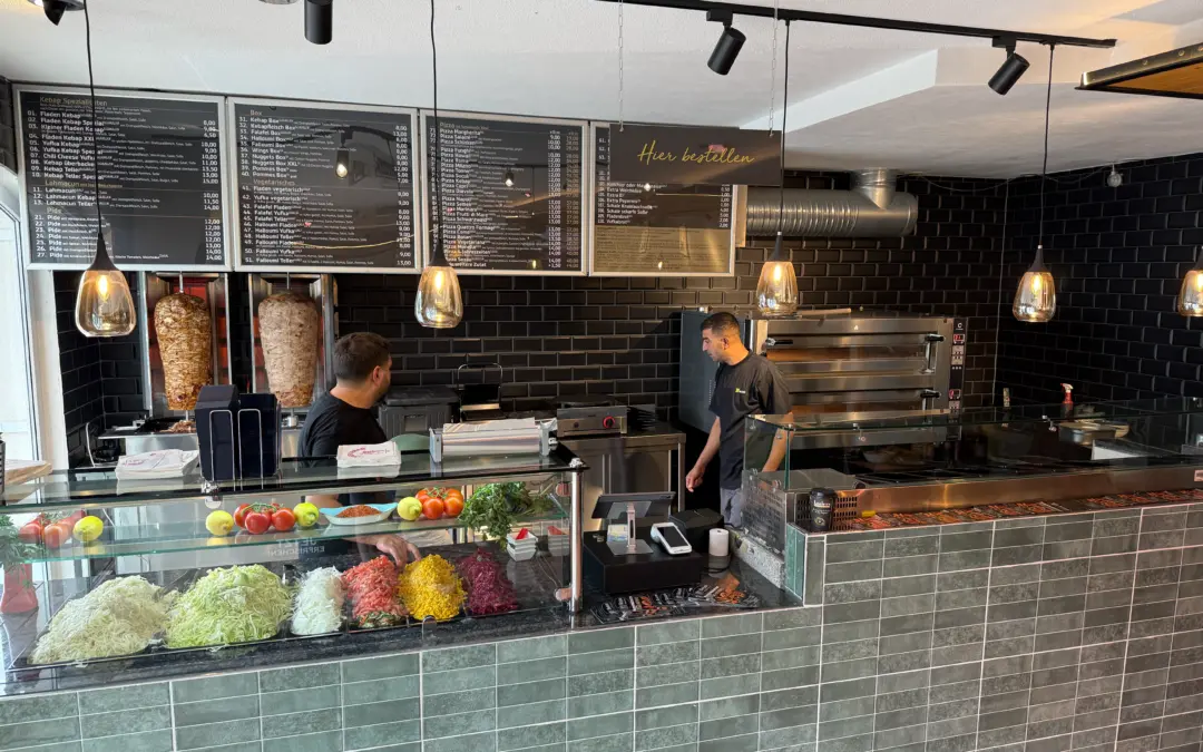 Kebab shop counter with vertical rotisseries, menu boards, and a glass display of vegetables as two staff work behind the counter.