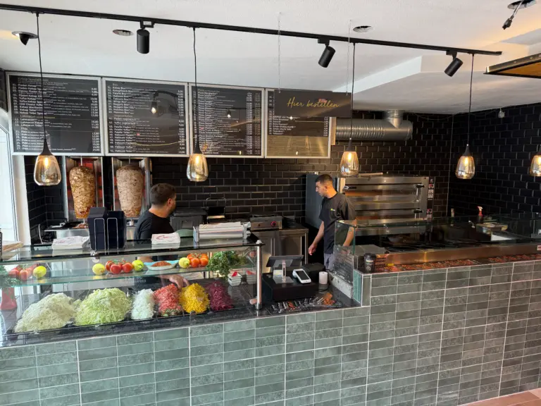 Kebab shop counter with vertical rotisseries, menu boards, and a glass display of vegetables as two staff work behind the counter.