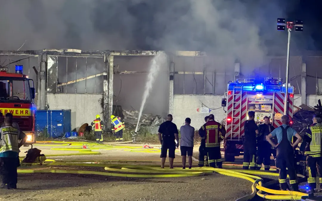 Nighttime scene of firefighters battling a warehouse blaze; smoke fills the air as hoses lay on the ground and the structure smolders.