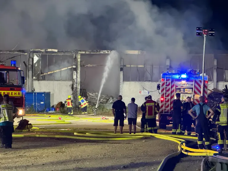 Nighttime scene of firefighters battling a warehouse blaze; smoke fills the air as hoses lay on the ground and the structure smolders.