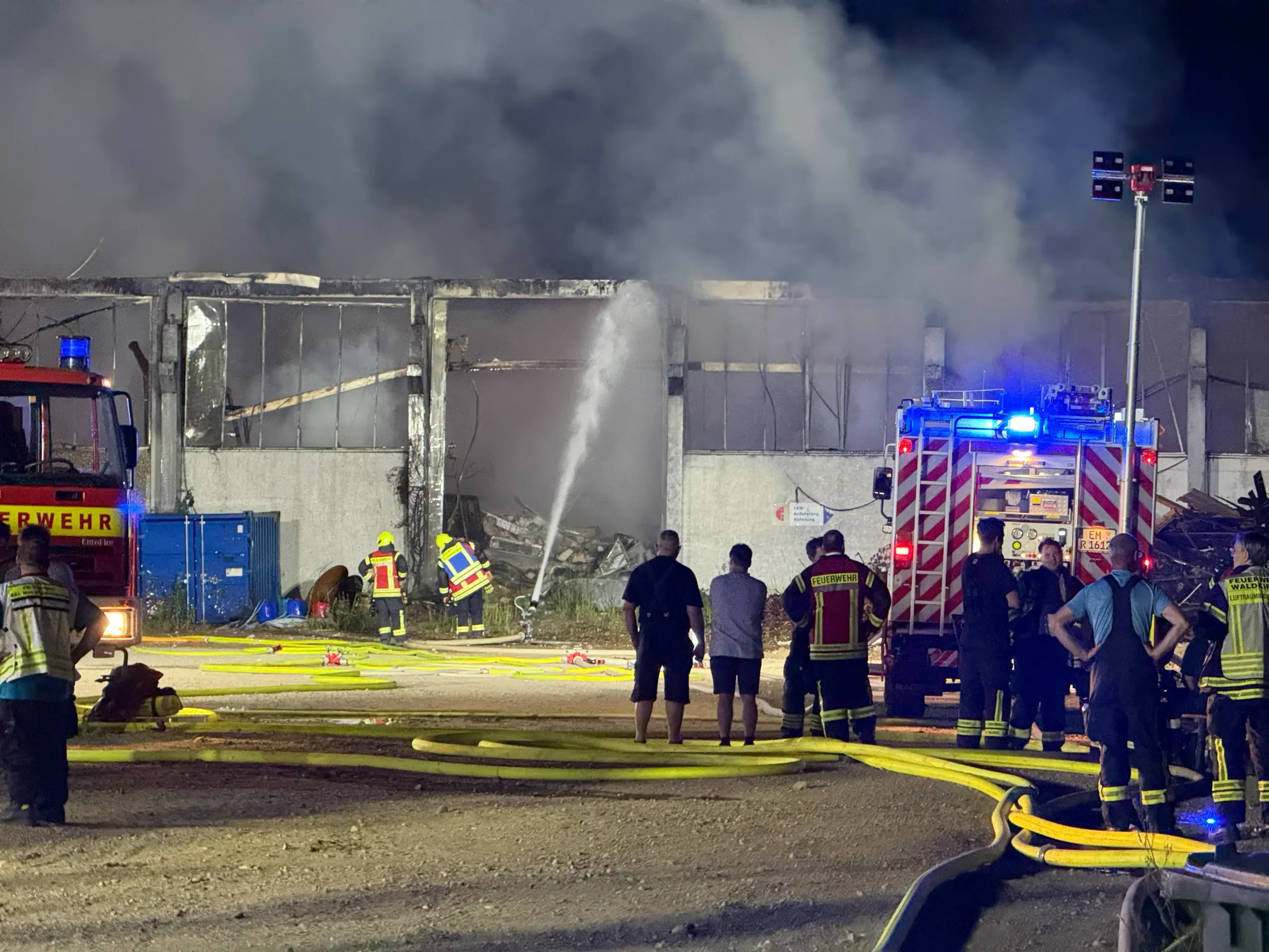 Nighttime scene of firefighters battling a warehouse blaze; smoke fills the air as hoses lay on the ground and the structure smolders.