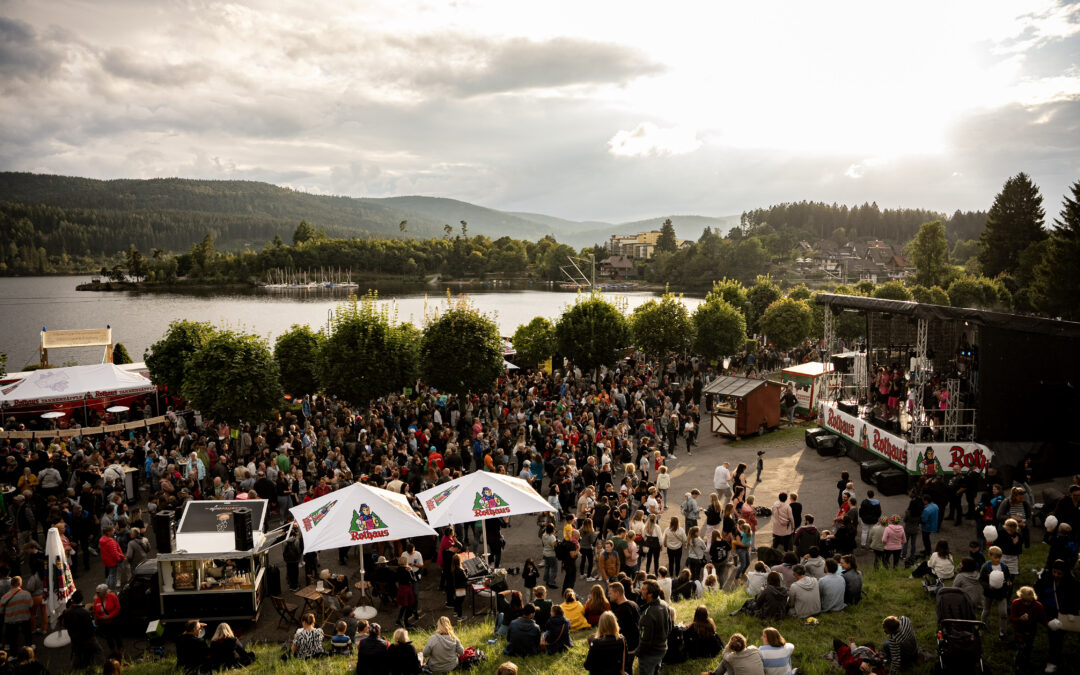 Large outdoor festival crowd gathered along a lake, with a stage on the right and white Rothaus tents in the foreground.