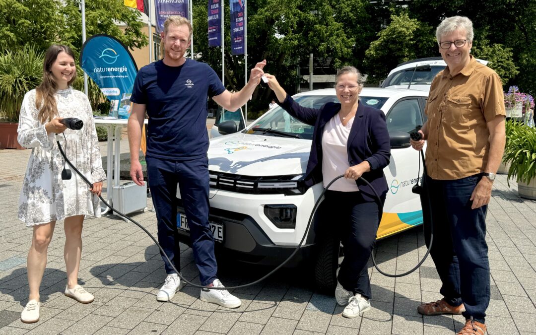 Four adults stand beside a white electric car at an outdoor event, holding charging cables and smiling at the camera.