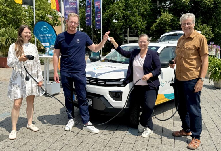 Four adults stand beside a white electric car at an outdoor event, holding charging cables and smiling at the camera.