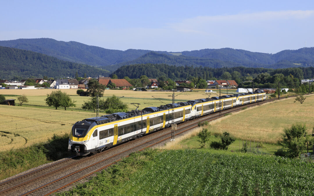 High-speed yellow-and-white passenger train traveling along tracks through a rural landscape with fields, trees, and distant hills under a blue sky.