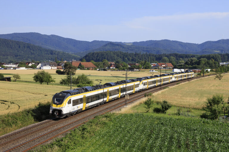 High-speed yellow-and-white passenger train traveling along tracks through a rural landscape with fields, trees, and distant hills under a blue sky.