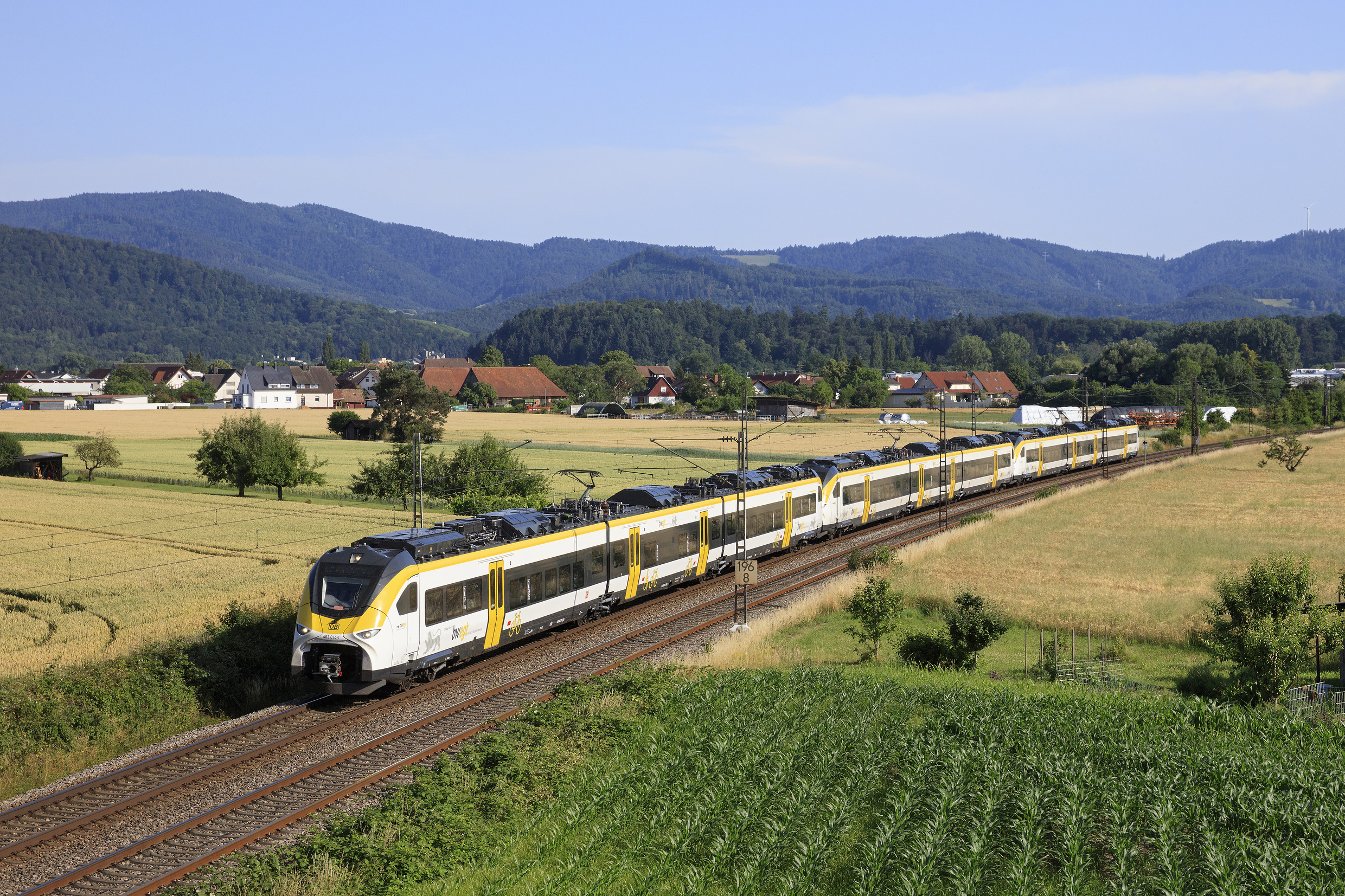 High-speed yellow-and-white passenger train traveling along tracks through a rural landscape with fields, trees, and distant hills under a blue sky.