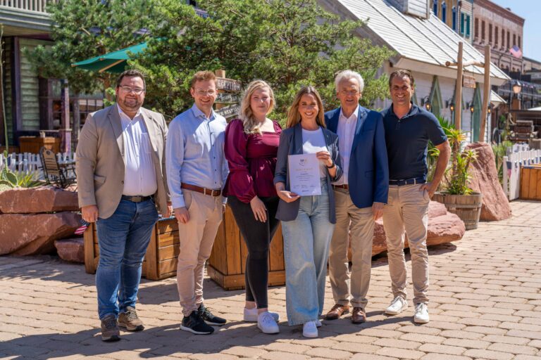 Six adults posing outdoors in a sunny street, one woman holding a certificate in front of shops and greenery?