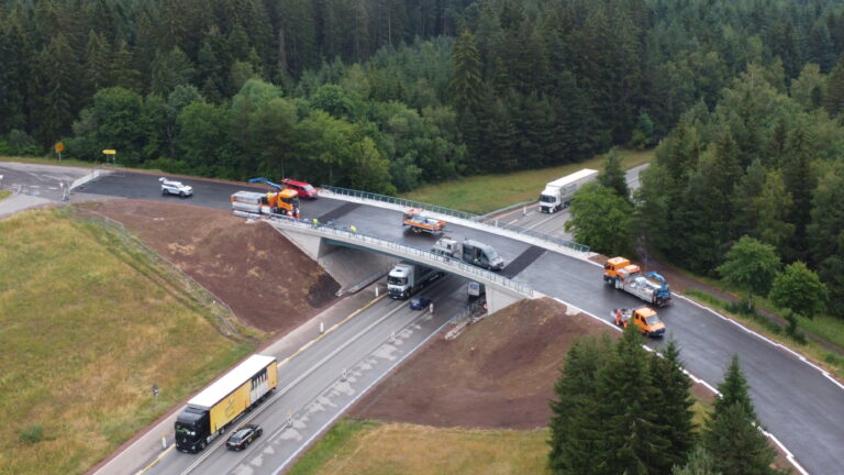 Aerial view of a road overpass under construction with trucks, workers, and construction equipment among a forested area.
