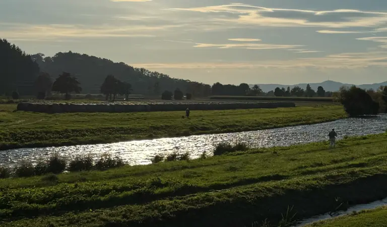 Riverbank with a person fishing near the water, fields and distant trees at sunset and rolling hills in the background.