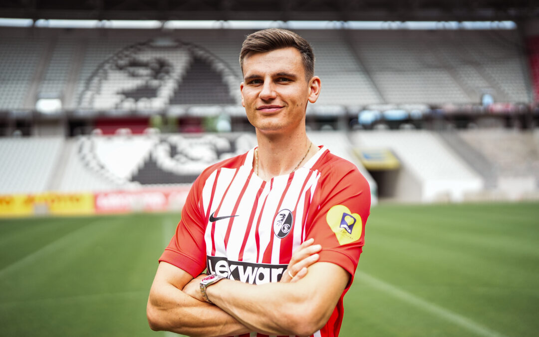 Soccer player in a red-and-white striped jersey stands with arms crossed on a green pitch, stadium seating behind him.