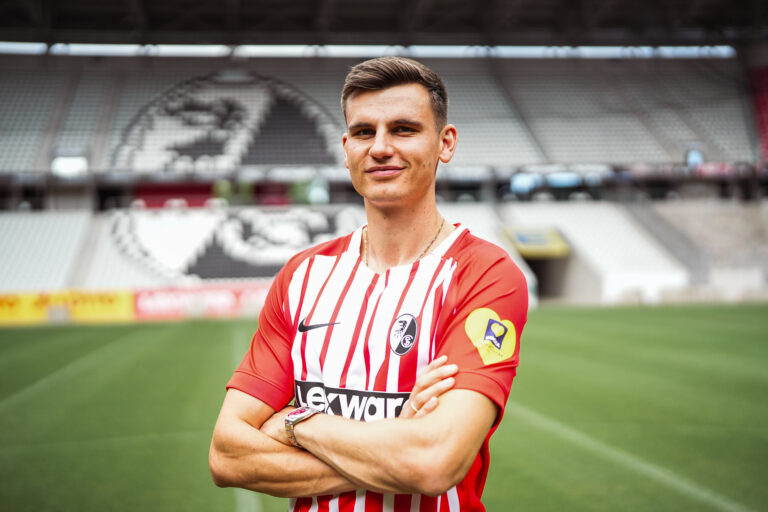 Soccer player in a red-and-white striped jersey stands with arms crossed on a green pitch, stadium seating behind him.