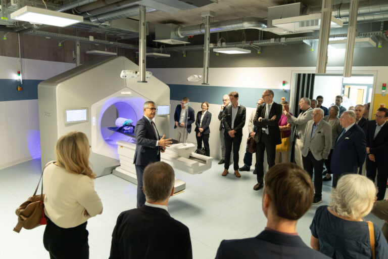 Professionals in business attire observe a large medical imaging machine in a clinical lab during a demonstration tour.