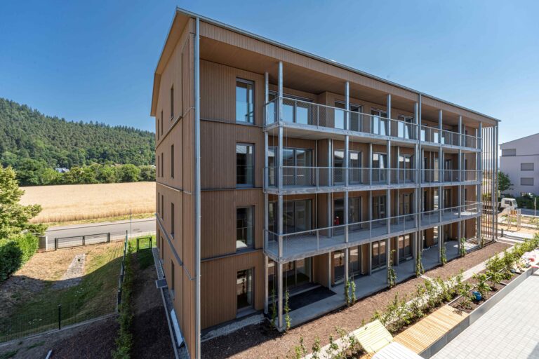 Four-story wooden apartment building with metal balconies and glass doors, set against a rural landscape with a wheat field and forested hills in the distance.