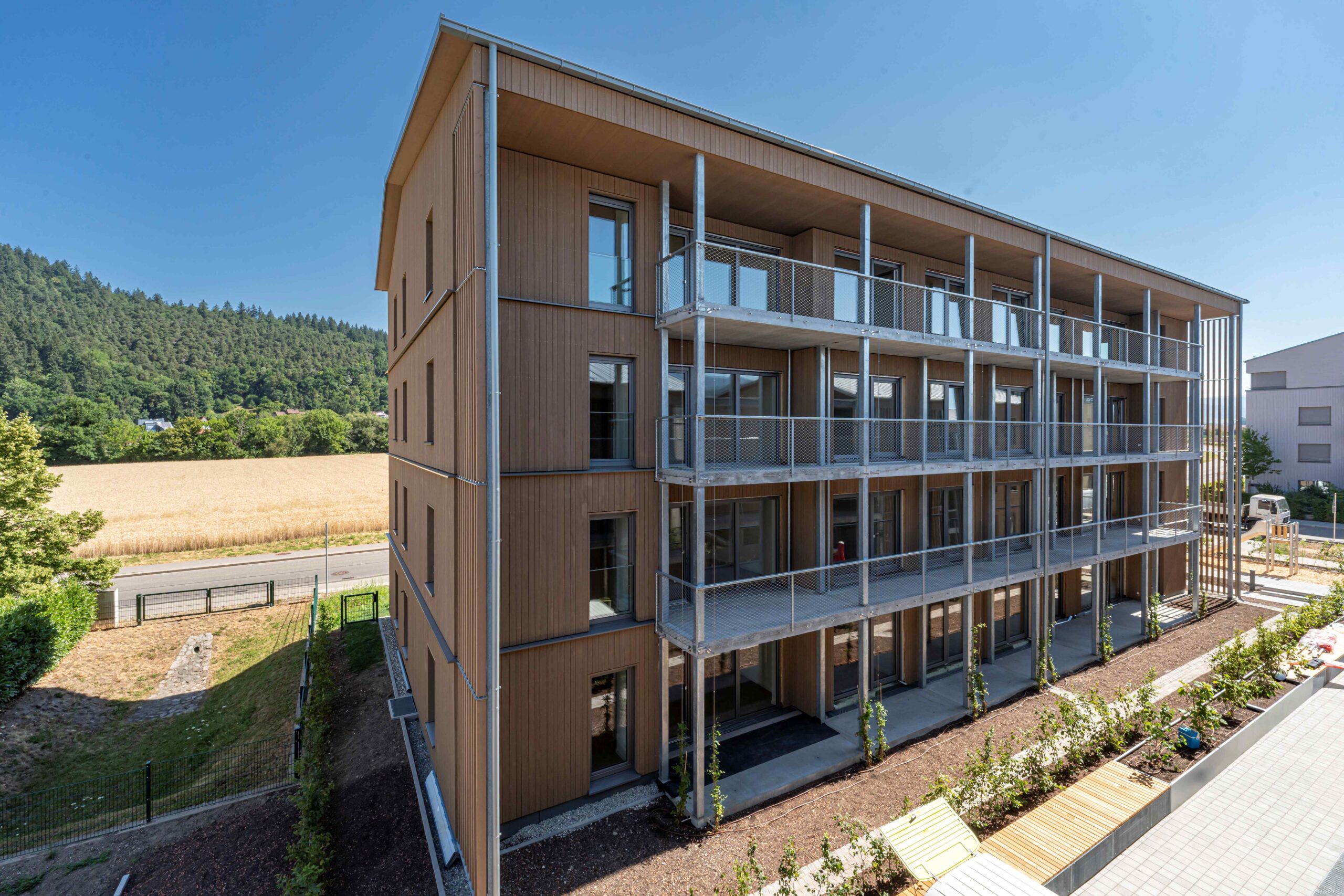 Four-story wooden apartment building with metal balconies and glass doors, set against a rural landscape with a wheat field and forested hills in the distance.