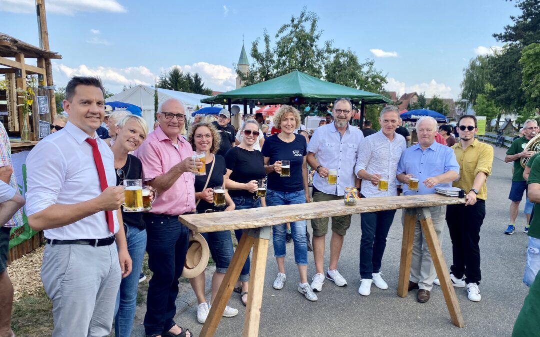 Group of people at an outdoor beer festival posing with beer mugs beside a wooden bar under a blue sky.