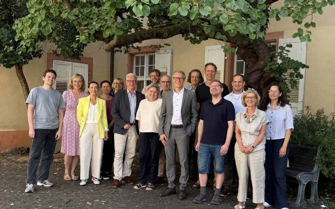 Group of about 20 adults posing for a photo outside a beige building with white shutters under a leafy tree.