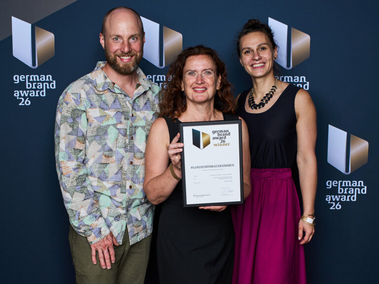 Three people posing with a framed German Brand Award certificate at an awards event.