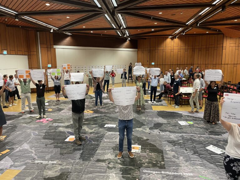 Group of people in a large, wood-paneled conference hall holding up large white sheets with handwritten notes, standing over a dark marble-like floor covered with printed names and maps.