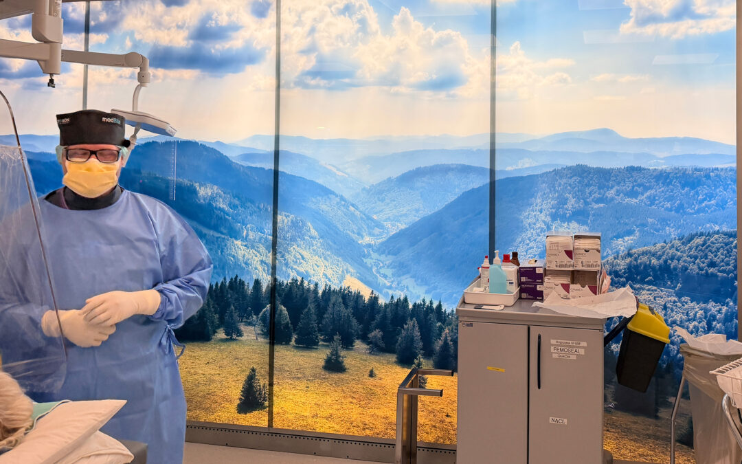 Medical professional in blue gown, yellow mask, and cap stands in a clinic with a panoramic mountain mural backdrop and medical supplies nearby.