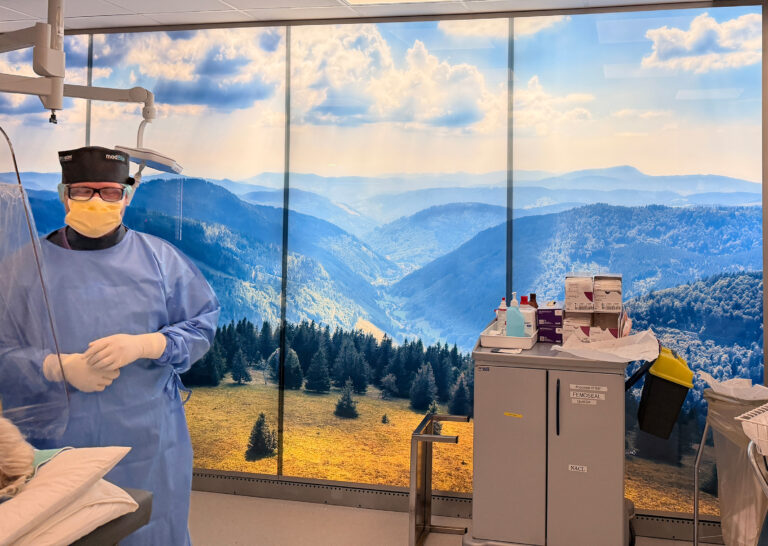 Medical professional in blue gown, yellow mask, and cap stands in a clinic with a panoramic mountain mural backdrop and medical supplies nearby.