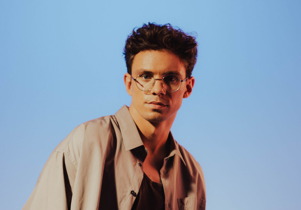 Young man with round glasses and tousled dark hair against a clear blue sky.
