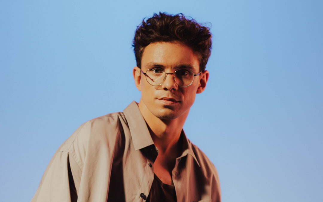 Young man with round glasses and tousled dark hair against a clear blue sky.