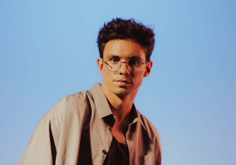Young man with round glasses and tousled dark hair against a clear blue sky.