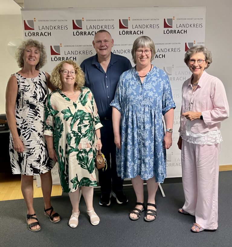 Five adults posing for a group photo in front of a backdrop with 'Landkreis Lörrach' logos; three women in patterned dresses on left and center, a man in a dark shirt, and a woman in a light purple outfit on the right.