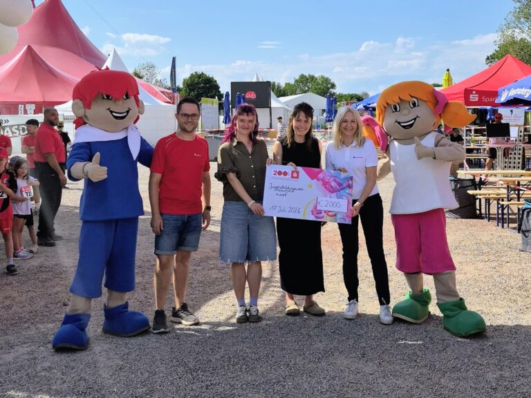Group of five adults standing with two large cartoon mascots at an outdoor festival, holding a big prize check together.