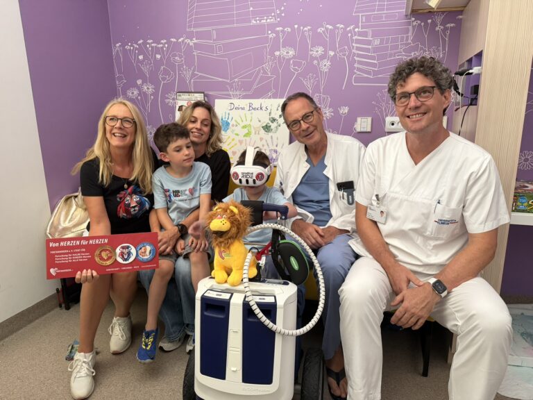 Group of smiling family and medical staff posing in a hospital room with purple wall art and a mobility scooter in the foreground.