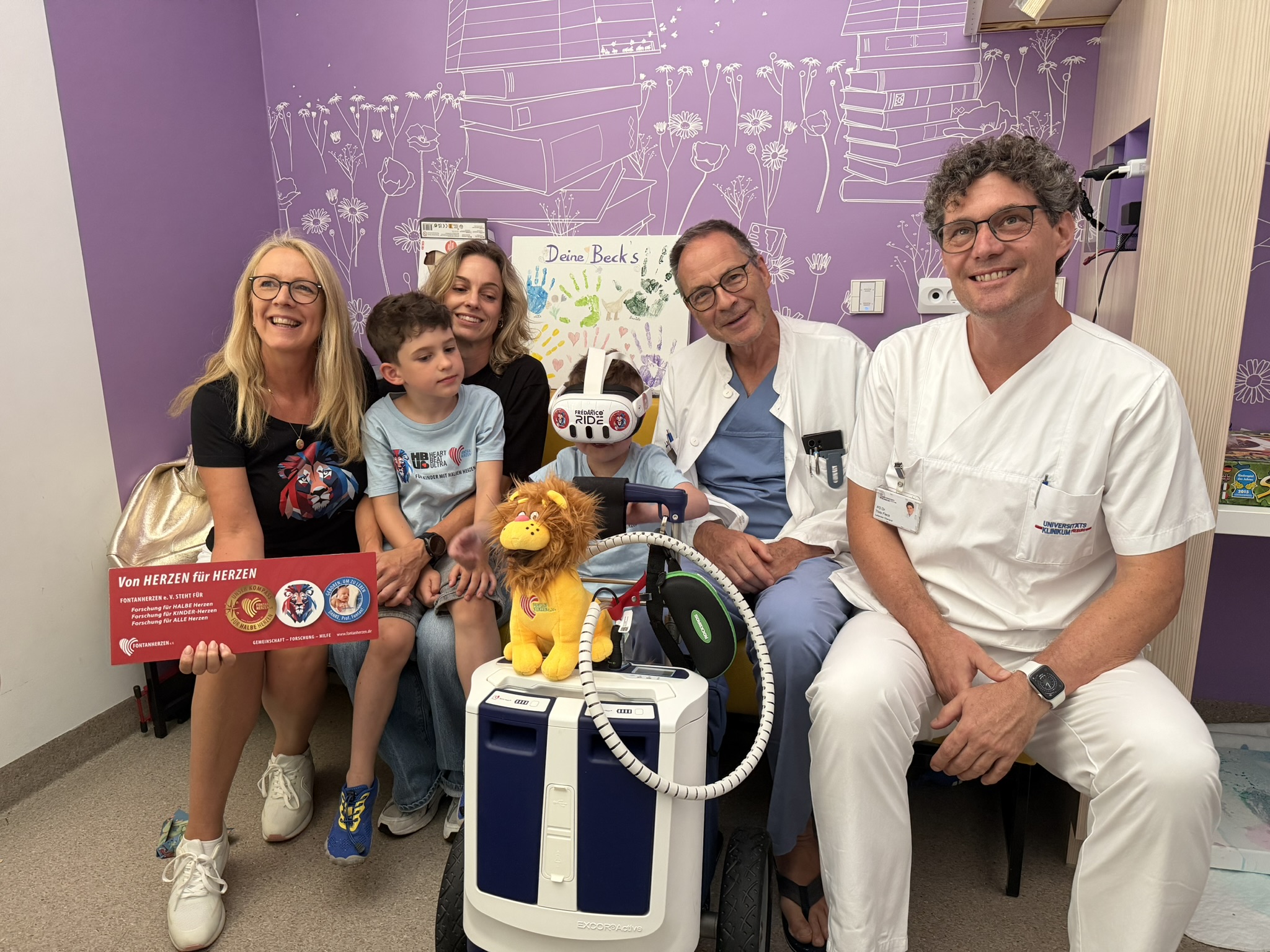 Group of smiling family and medical staff posing in a hospital room with purple wall art and a mobility scooter in the foreground.