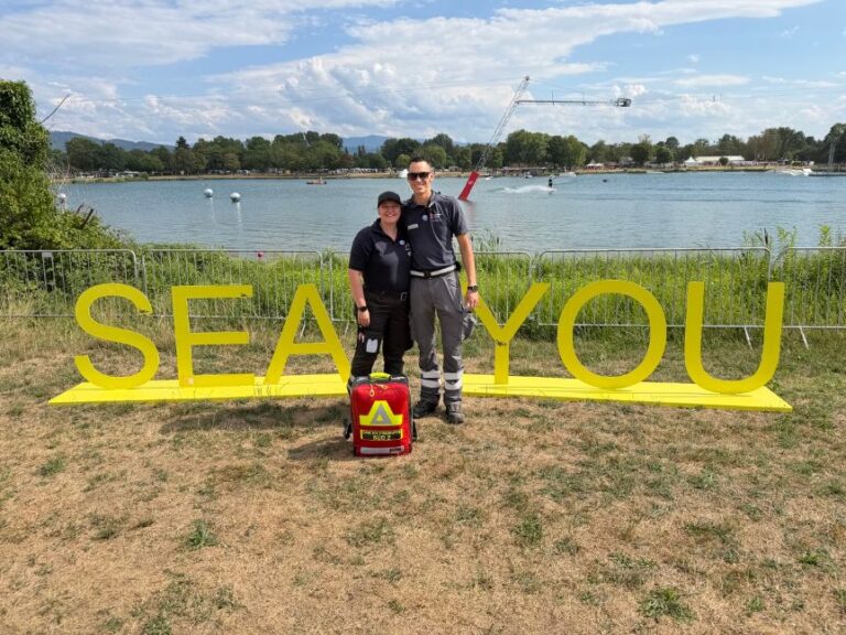 Two people in dark uniforms stand side by side by a lakeside, smiling, with a bright yellow 'SEA YOU' sign behind them and a red first-aid bag on the ground in front of them.