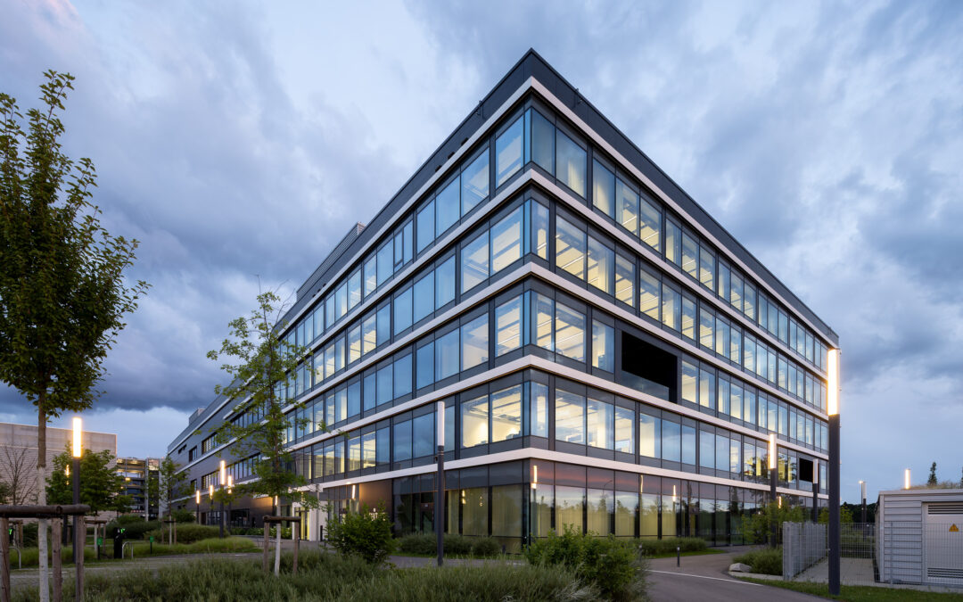 Modern glass-fronted office building at dusk with illuminated interiors and surrounding landscaping in the foreground.